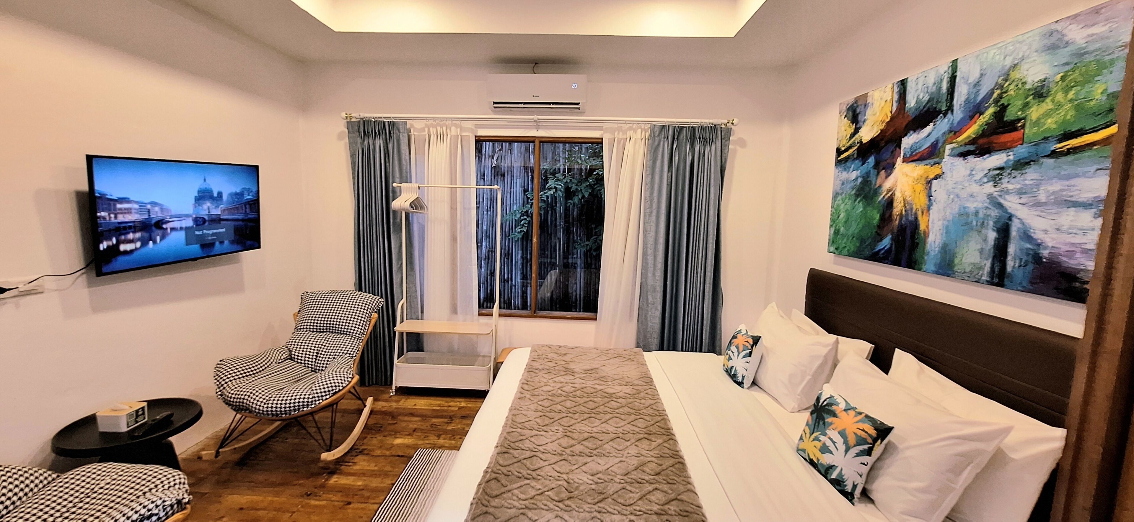 Una habitación de hotel con cama, televisión, silla, ventana con cortinas y un cuadro.