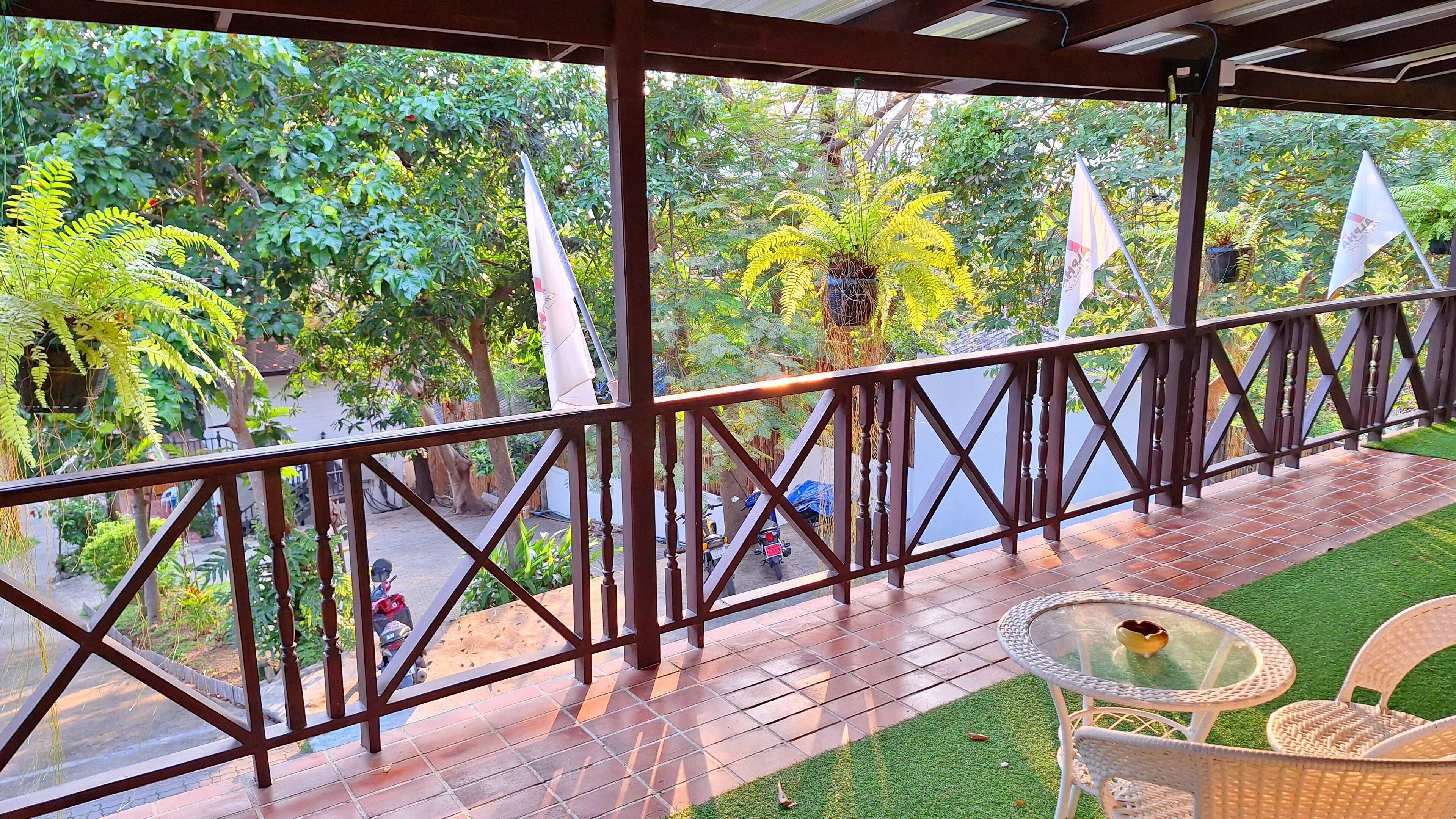 A covered outdoor area with a wooden railing, a small table with chairs, and a view of a garden with trees and a parked motorcycle.