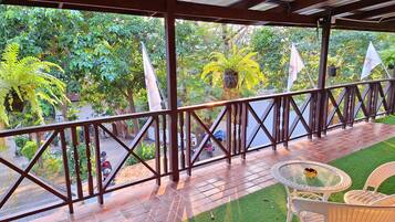 A covered outdoor area with a wooden railing, a small table with chairs, and a view of a garden with trees and a parked motorcycle.