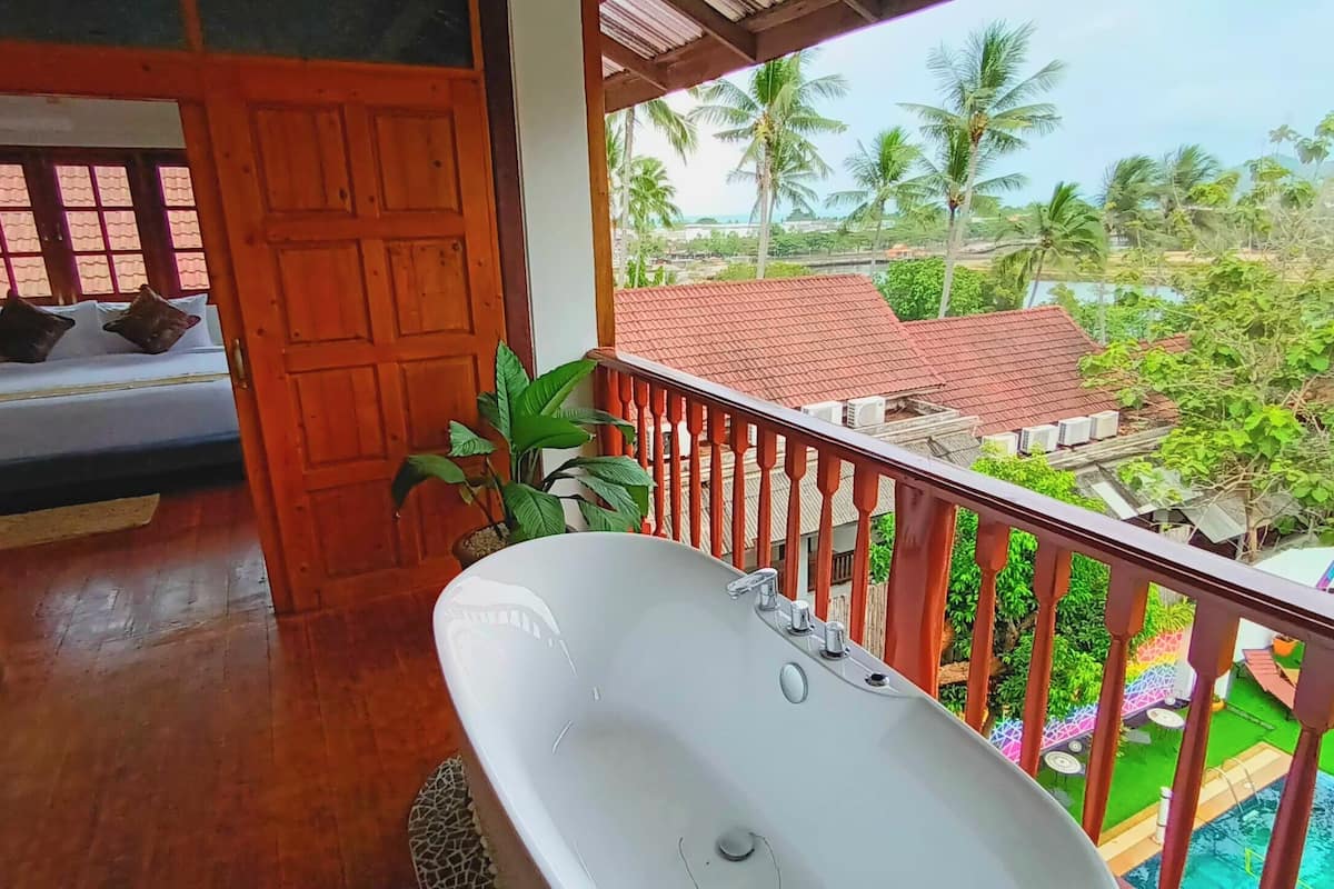 A balcony with a white bathtub, wooden door, and a view of a pool and palm trees.
