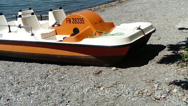 En båt med registreringsnummer FN 28335 ligger vid en strand med småsten, intill ett stilla vatten.