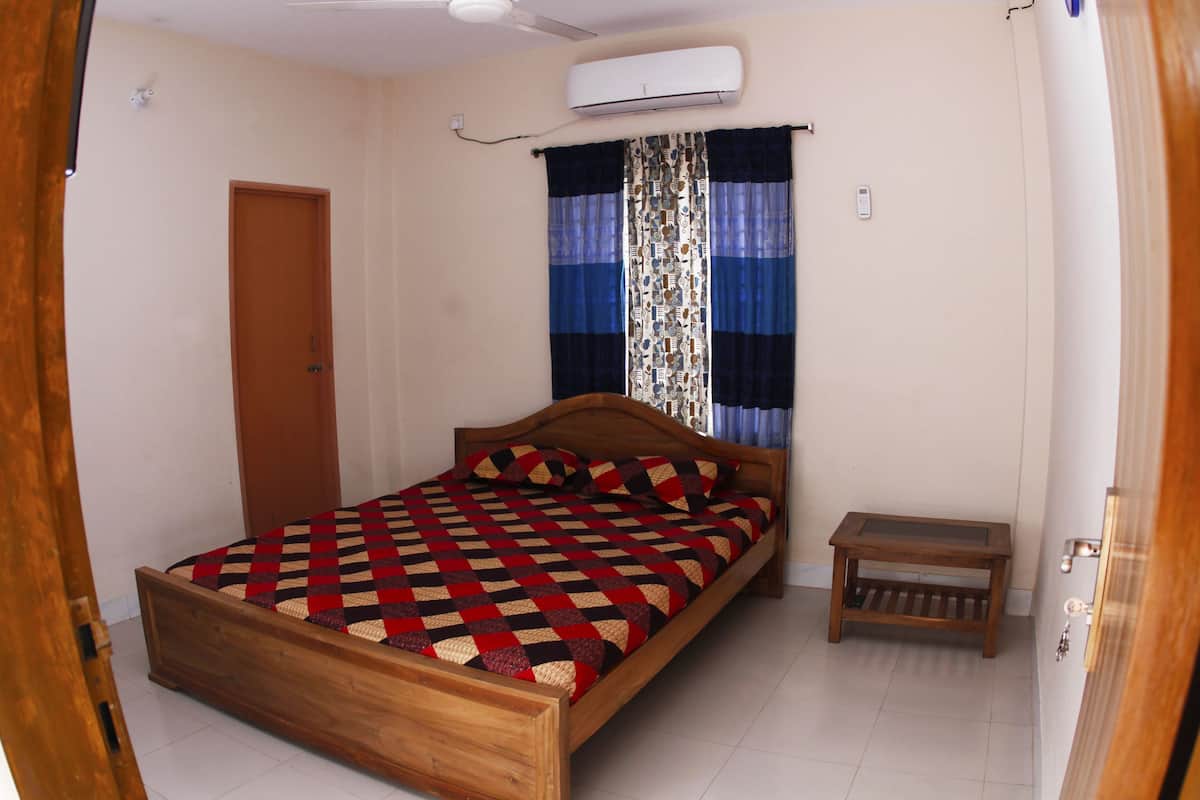 A bedroom with a wooden bed, a small bedside table, a door, and a window with patterned curtains.