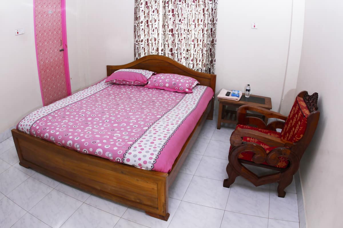 A neatly made bed with a patterned bedspread and pillows, a wooden nightstand with a water bottle, and a traditional wooden chair with a red cushion.