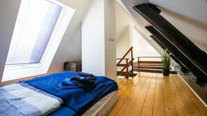 Un dormitorio con tragaluz, suelo de madera, una cama con ropa de cama azul, una escalera y una planta en maceta.