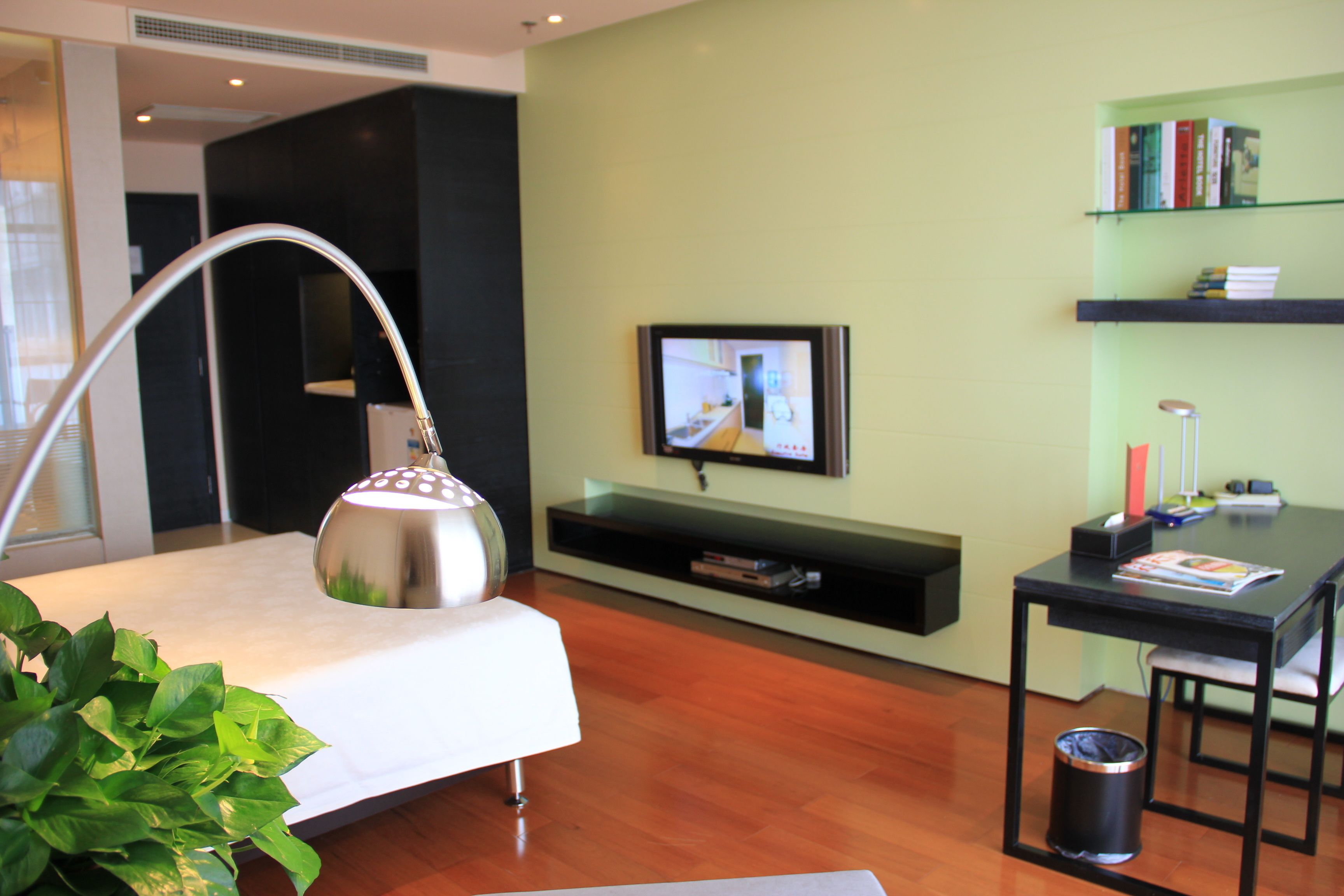 A modern hotel room with a bed, a desk, a TV, a bookshelf, and a potted plant.