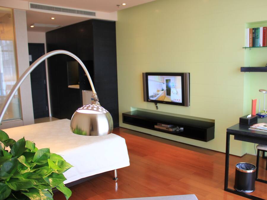 A modern hotel room with a bed, a desk, a TV, a bookshelf, and a potted plant.
