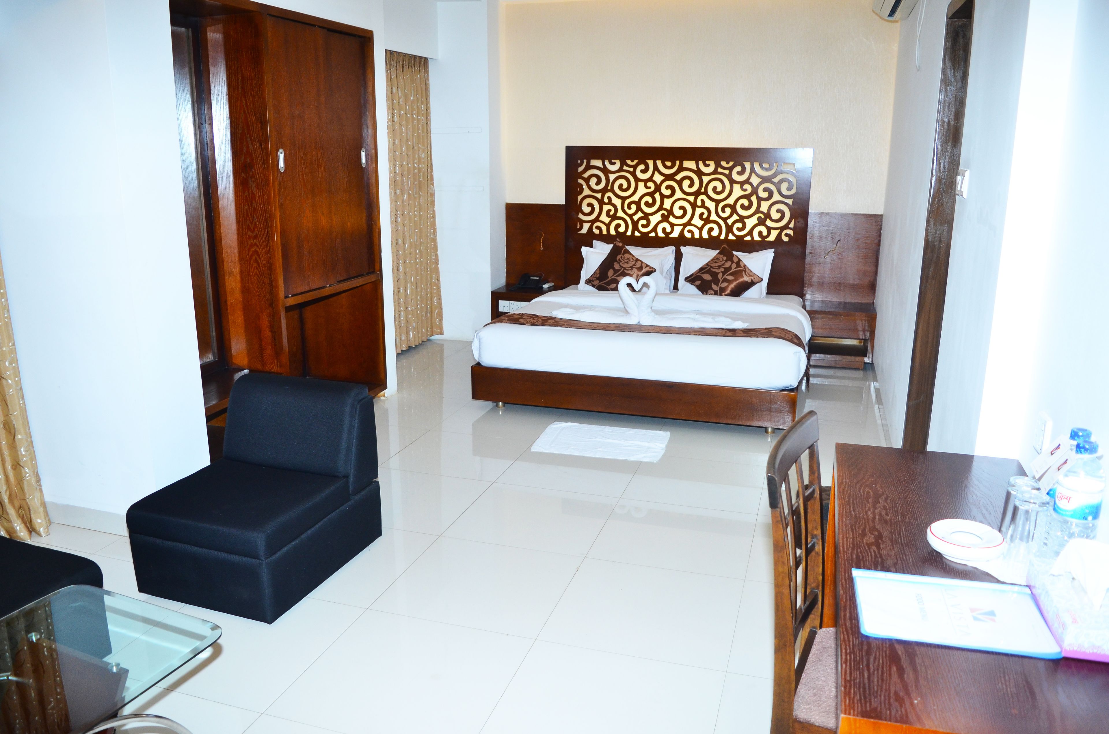 A hotel room with a large bed, a wooden wardrobe, a small black sofa, a glass table, and a wooden desk with a chair.