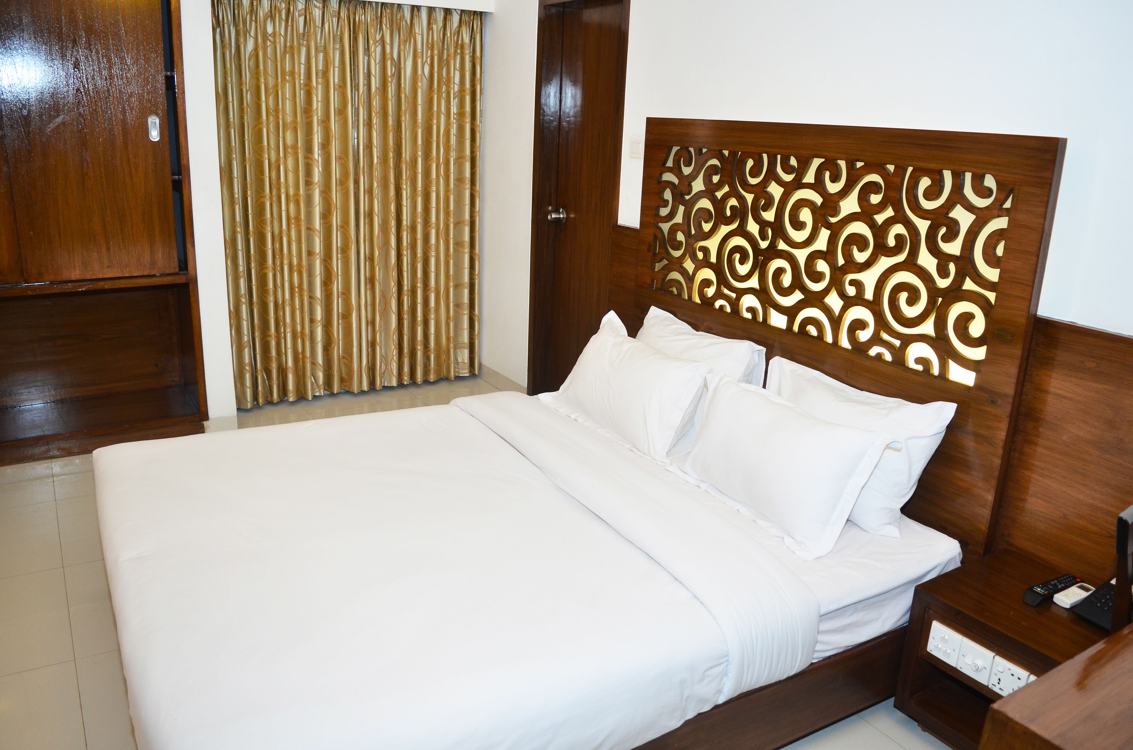 A neatly made bed with white linens, a wooden headboard with intricate carvings, and a bedside table with a remote control.