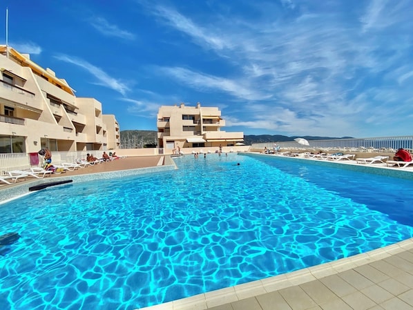 A swimming pool with clear blue water, surrounded by buildings with balconies and a clear sky.