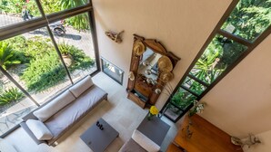 Una sala de estar moderna con un sofá, una mesa de centro y una mesita de noche de madera. Hay un espejo grande en la pared y una ventana con vistas a un jardín.