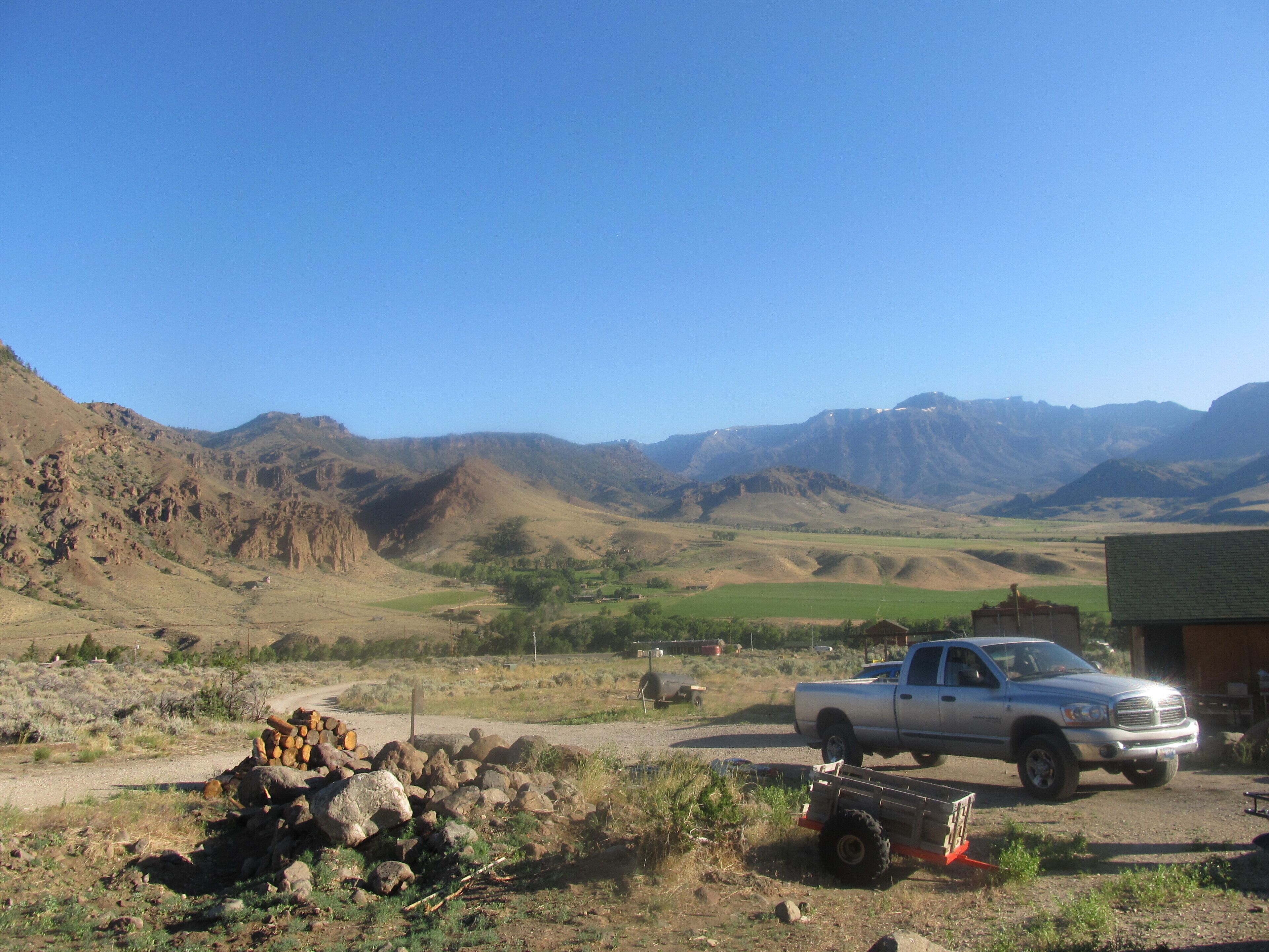 A rural landscape with a silver pickup truck parked near a building, surrounded by mountains and a clear sky.