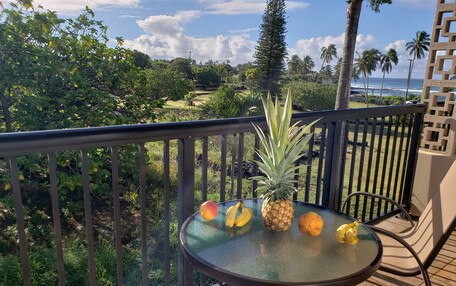 Un balcón con una mesa de vidrio que sostiene frutas, una silla y vista al océano y palmeras.