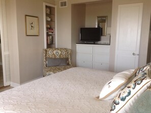A bedroom with a bed, a television on a dresser, a chair, and a bookshelf.