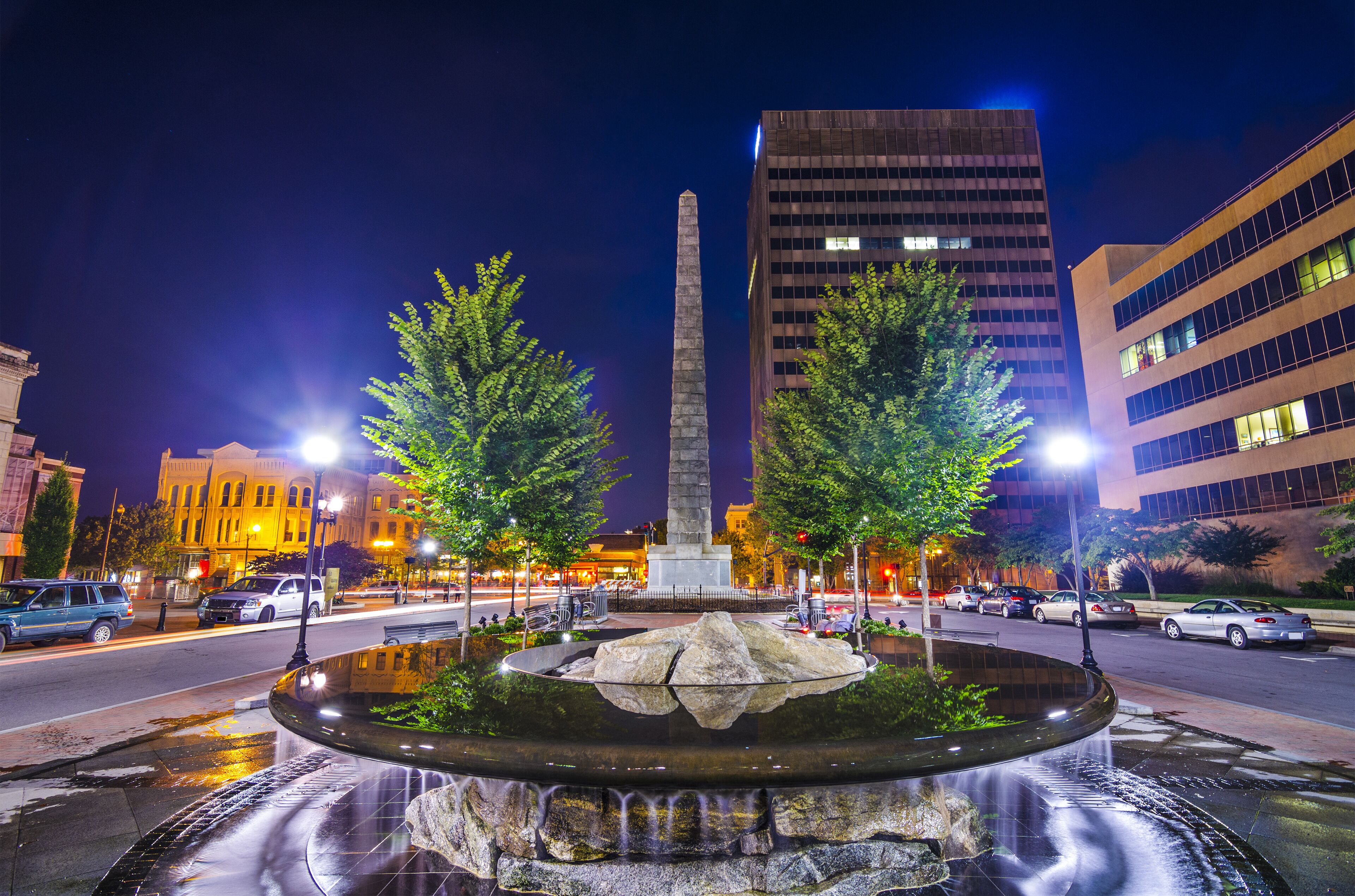 Une scène urbaine nocturne avec un monument central, une fontaine et des bâtiments modernes environnants.