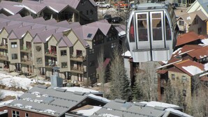 Une télécabine se déplace au-dessus d’un quartier résidentiel, avec des immeubles à plusieurs étages et des toits enneigés.
