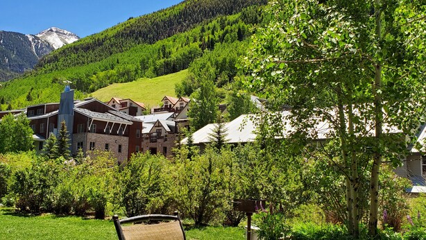 Une table ronde en métal, accompagnée d’une chaise en osier, est placée dans un espace verdoyant, offrant une vue sur une montagne et des bâtiments en arrière-plan.