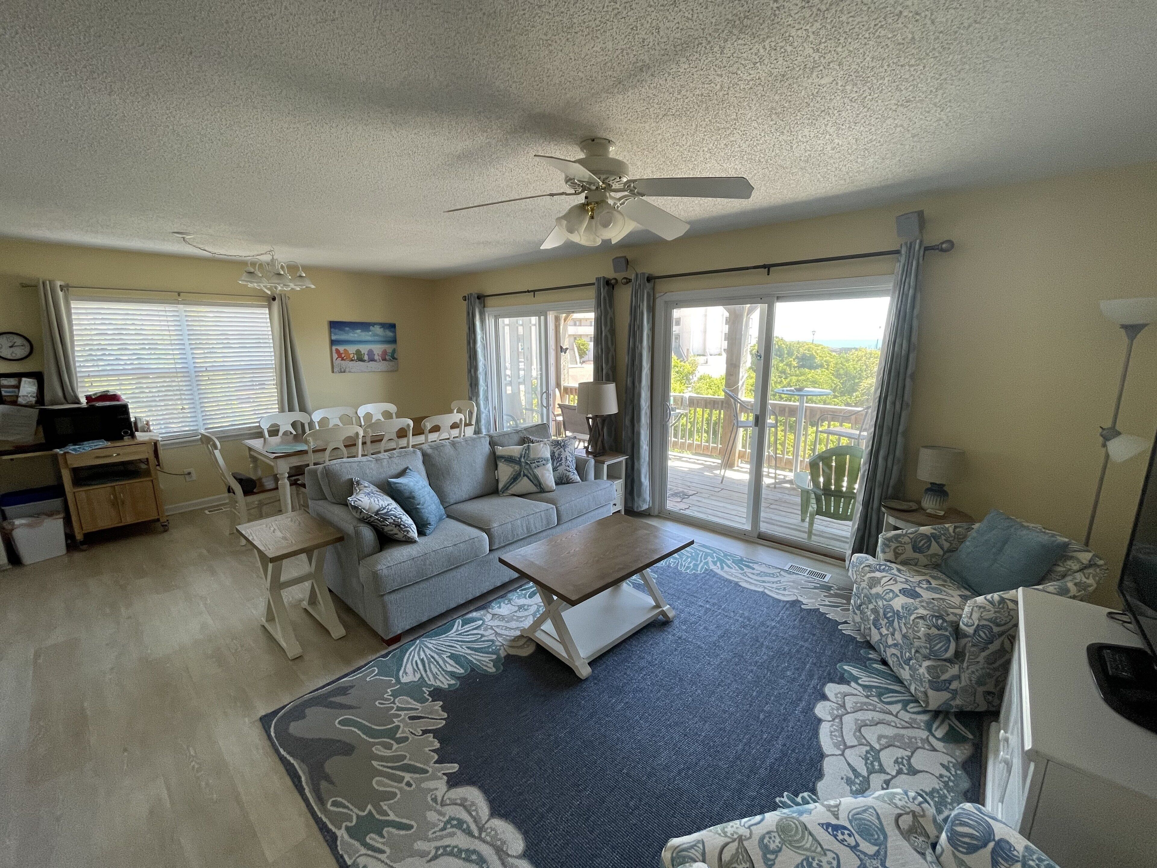 A living room with a sofa, coffee table, and chairs. There is a ceiling fan, a window with blinds, and a view of the outdoors.