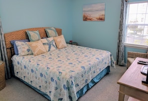 A bedroom with a bed, a nightstand, a wicker chair, and a window with curtains. - 2nd ROW EASY beach access 1/2 block Ocean View + EV charger (Emerald Isle)