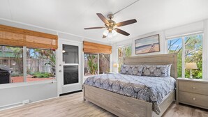 Un dormitorio con cama, ventilador de techo, ventana con vista al jardín trasero y mesita de noche con lámpara.