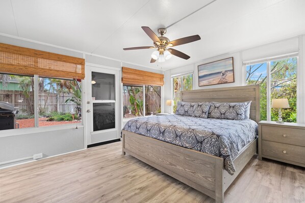 A bedroom with a bed, a ceiling fan, a window with a view of the backyard, and a nightstand with a lamp.