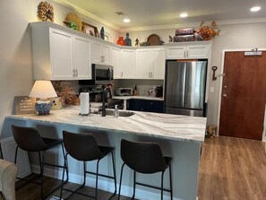 Una cocina moderna con armarios blancos, una isla de mármol con taburetes altos, electrodomésticos de acero inoxidable y objetos decorativos en los estantes.
