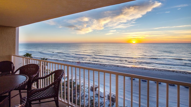 Un balcon avec des fauteuils en osier et une table donnant sur l’océan au coucher du soleil.