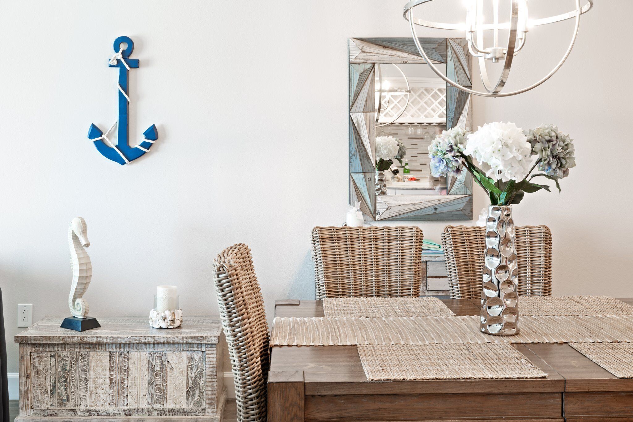 Uma área de refeições com uma mesa de madeira, cadeiras de vime, um vaso decorativo com flores e um adorno de âncora azul na parede.