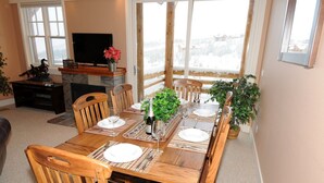 A dining area with a wooden table set for four, surrounded by wooden chairs. A television is mounted on the wall, and there are windows providing a view of a snowy landscape. - Casa Moonlight Two - Ideal Rental for Summer Activities and Trips to Yellowstone (Big Sky)