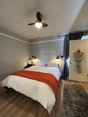 Un dormitorio con cama, mesitas de noche, ventilador de techo, bata colgada y una puerta.