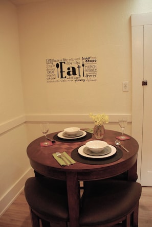 A dining area with a round table set for two, a wall decal with food-related words, and a vase with flowers. - Waterfront Historic Bungalow One• Ferry to SF (Vallejo)