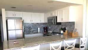 A modern kitchen with white cabinets, a stainless steel refrigerator, a microwave, and a granite countertop. There are four white bar stools with black cushions. - Beachfront...South Corner 3 Bedroom With Spectacular Views (South Padre Island)