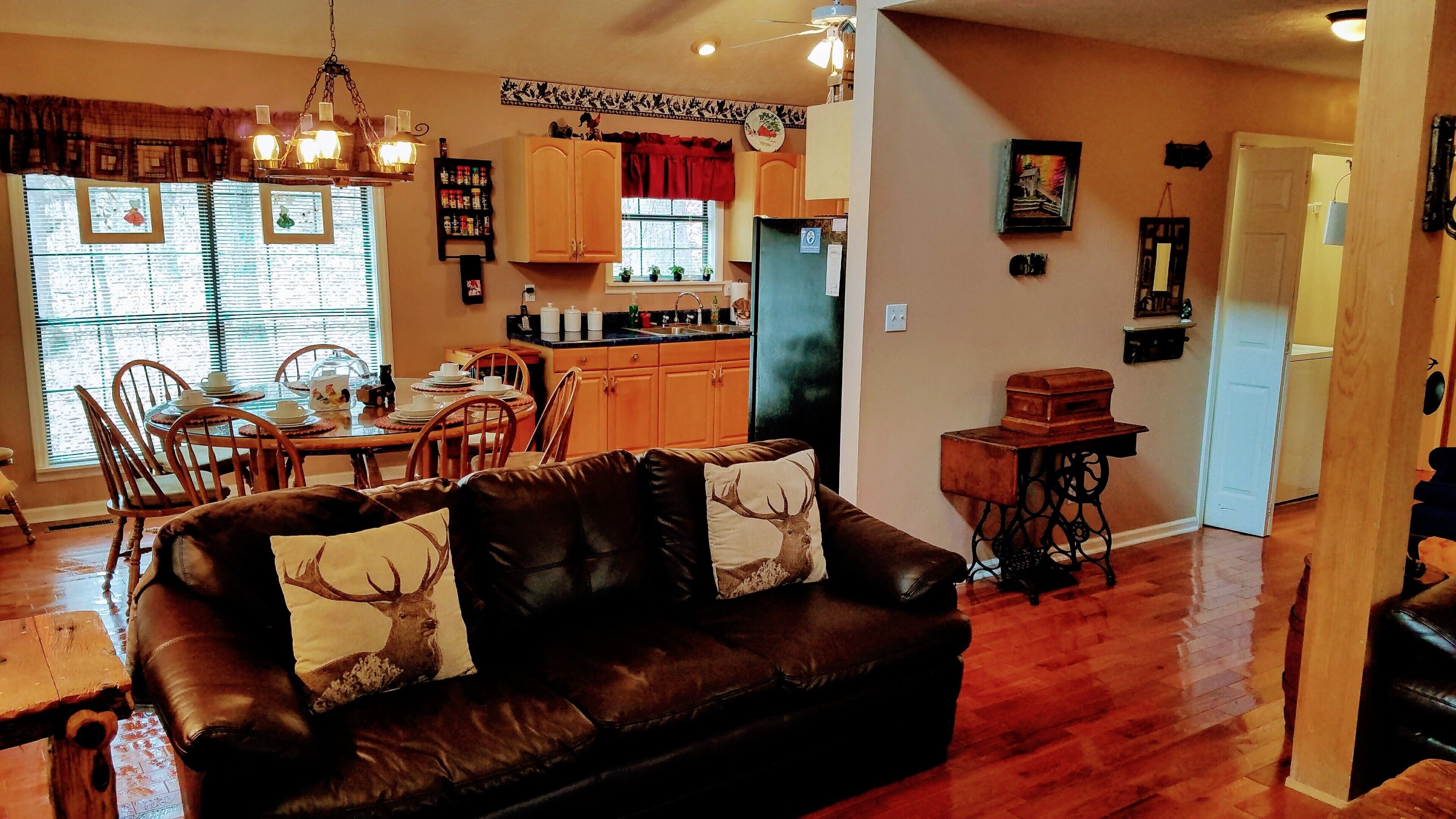 A cozy living room with a brown leather sofa, a dining area with wooden chairs and a table, and a kitchen with wooden cabinets and a refrigerator.