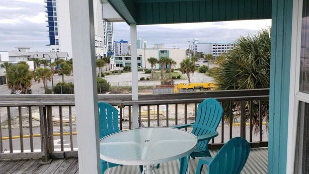 A balcony with a glass table and turquoise chairs, overlooking a street with palm trees and buildings. - Private Upscale Condo in Myrtle w/ Ocean View Across from Boardwalk w/ Huge Deck (Myrtle Beach)