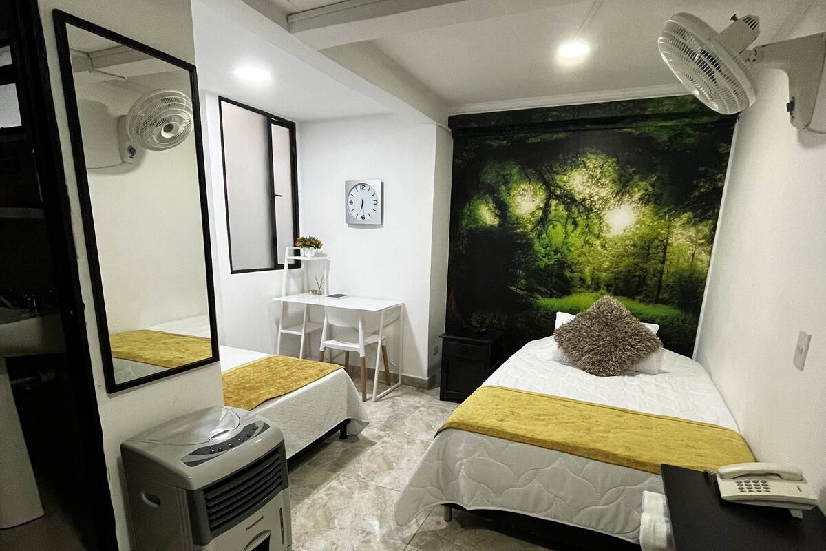 Habitación de hotel con cama, mesita de noche, espejo, calefactor y ventana grande con vista al bosque.