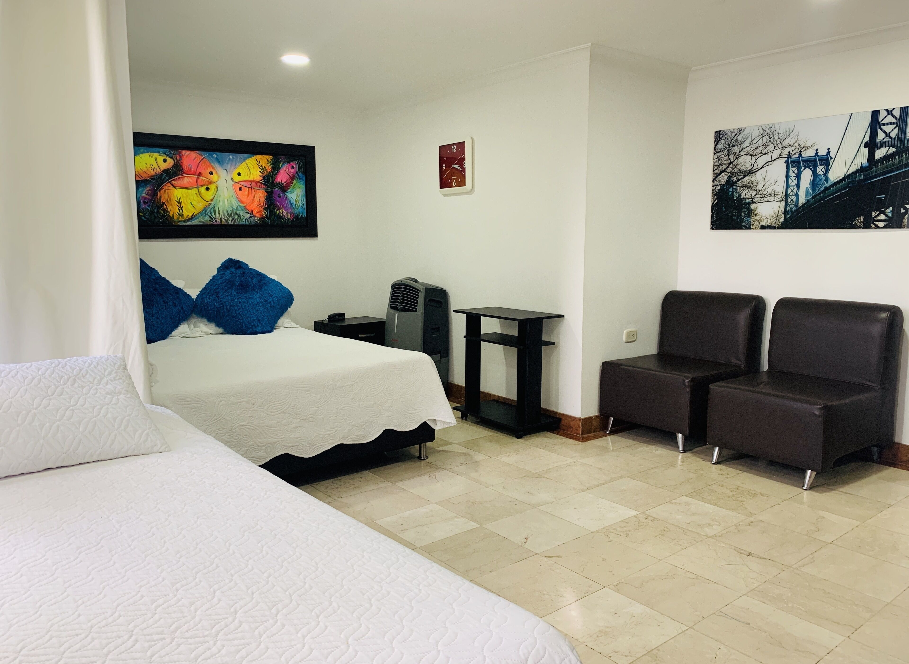 A hotel room with a bed, a desk, two chairs, a heater, a clock, and a painting of a bridge.