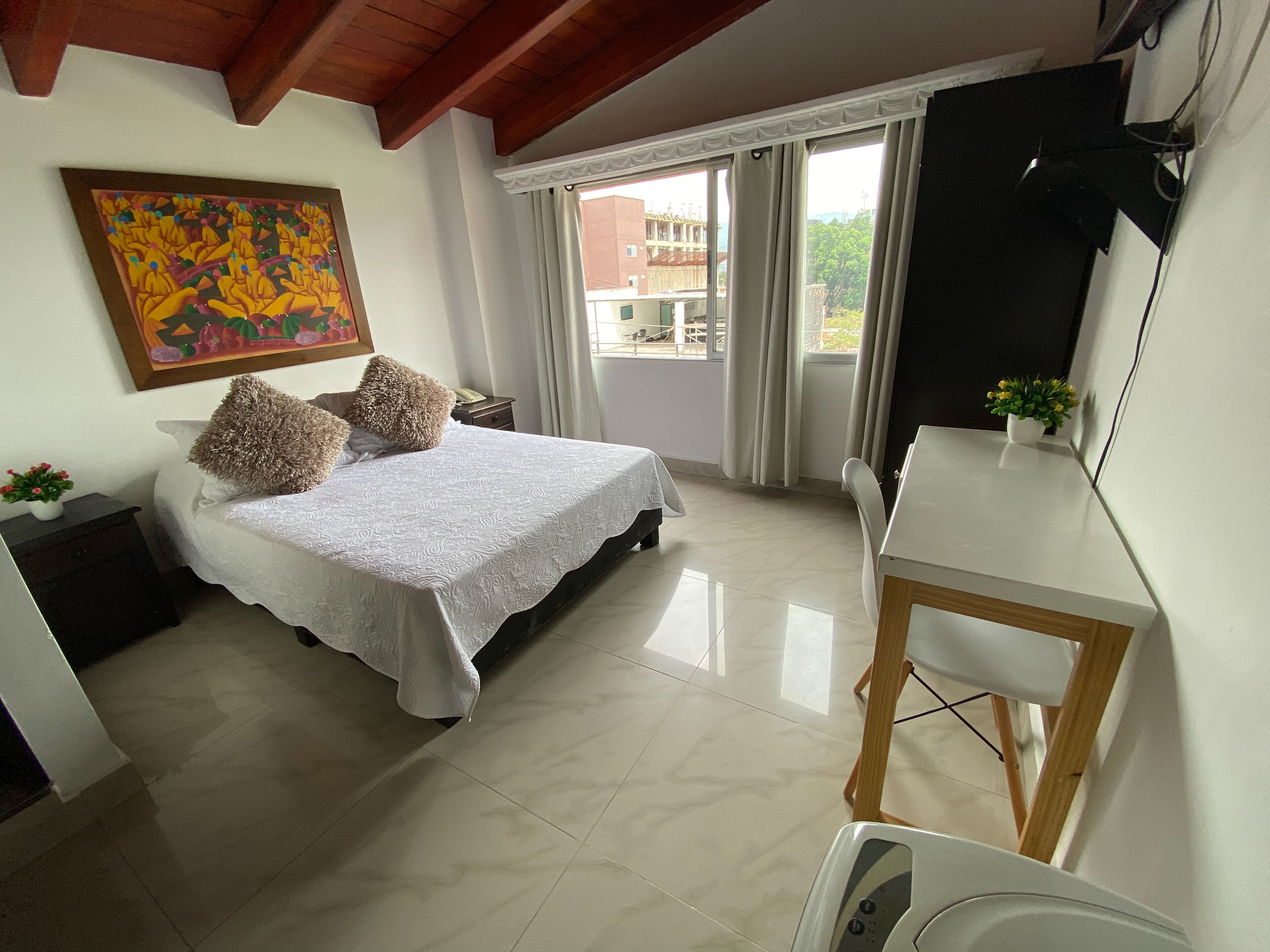 A bedroom with a bed, a desk, a chair, a painting, and a balcony with curtains.
