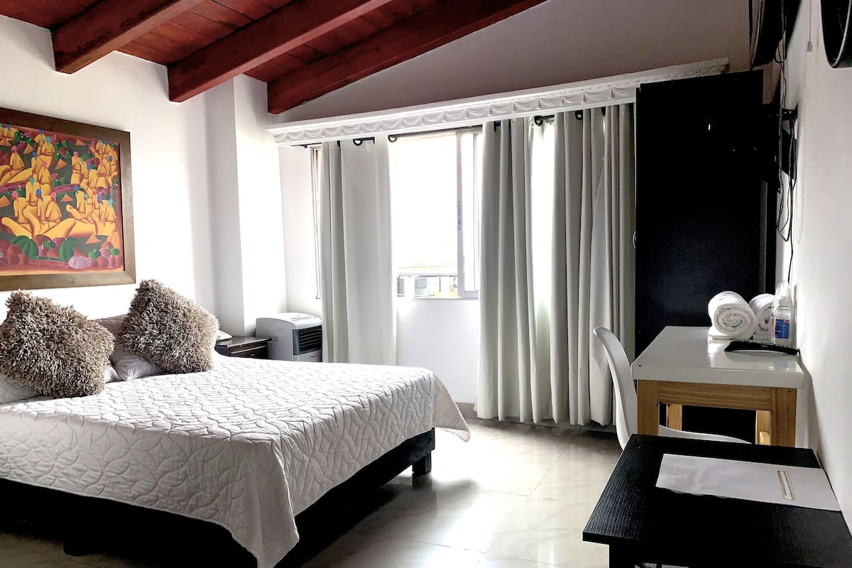 Una habitación de hotel con cama, escritorio, silla, un cuadro y ventana con cortinas.