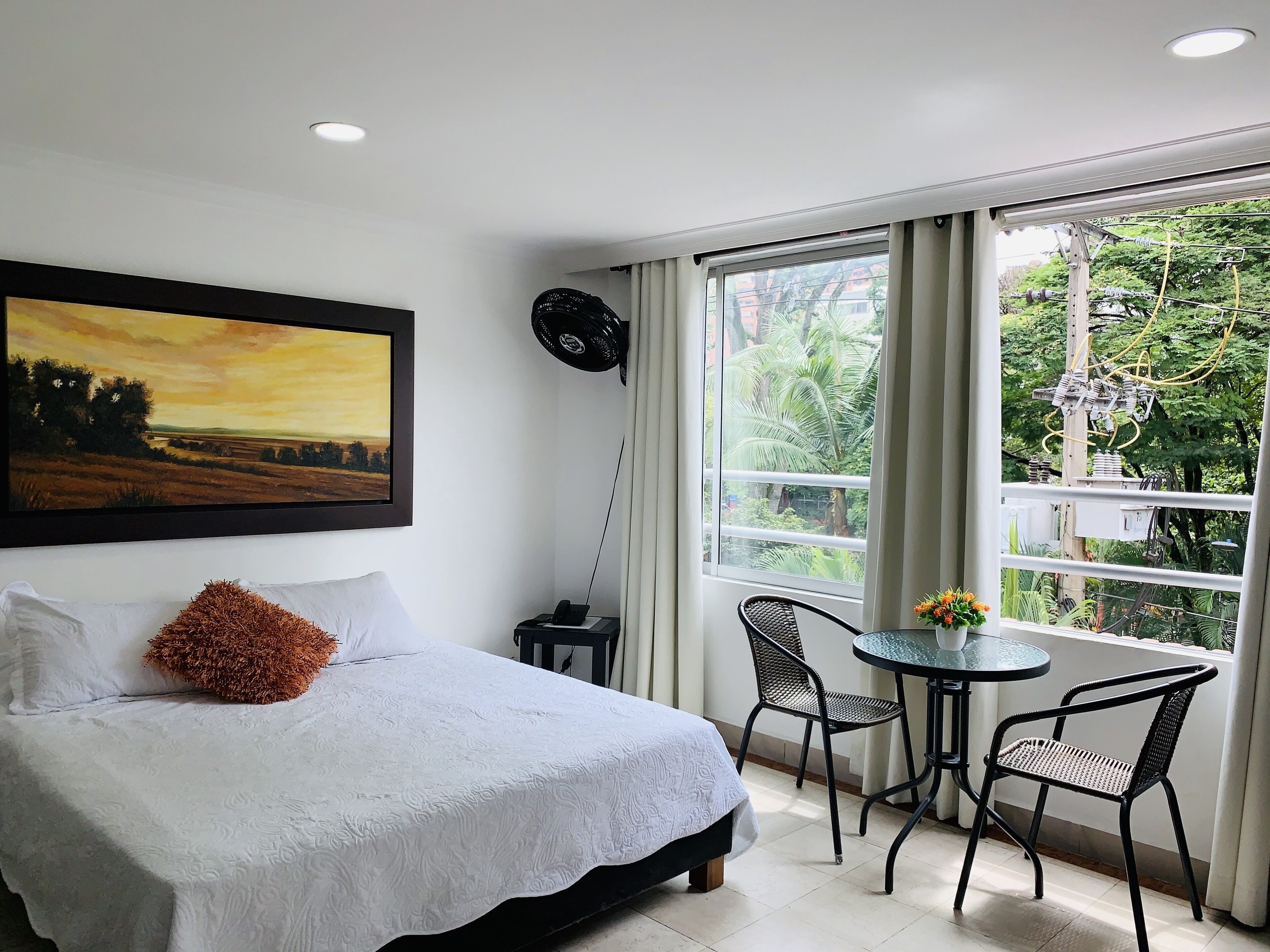A bedroom with a bed, a painting, a window with curtains, a table with chairs, and a view of greenery outside.