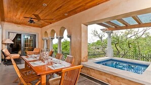 Un área de comedor al aire libre con mesa y sillas de madera, ventilador de techo y vista a una piscina.