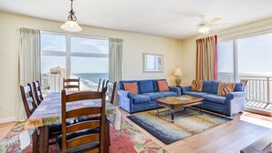 A living room with a view of the ocean, a dining area with a table and chairs, and a blue sofa with colorful pillows. - HUGE Seasonal Discounts & Promotions, Beachfront HUGE End Unit, FREE BeachCHAIRS (Panama City Beach)