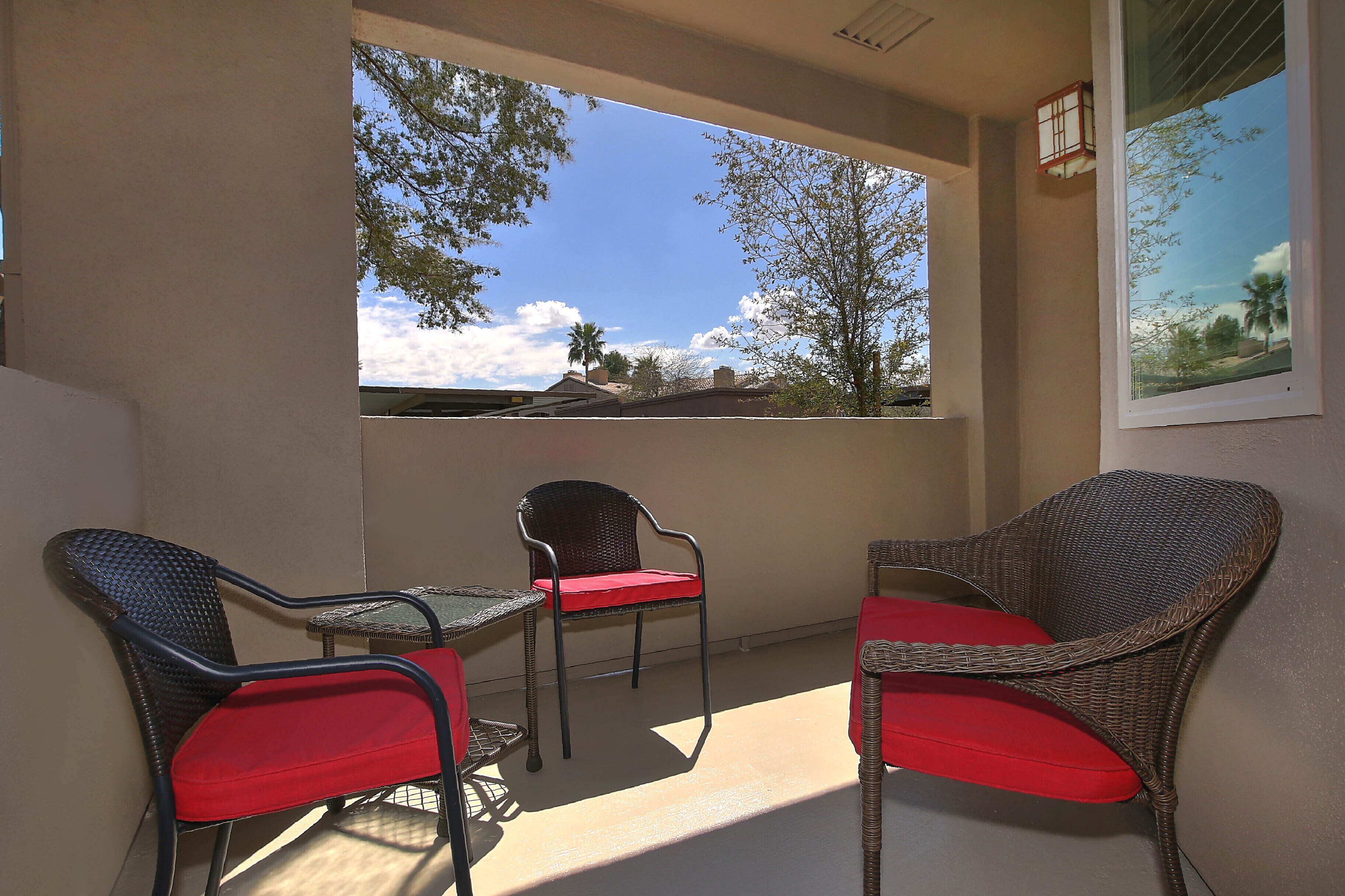 A balcony with wicker chairs and a table, a view of trees and a clear sky, and a framed picture on the wall.