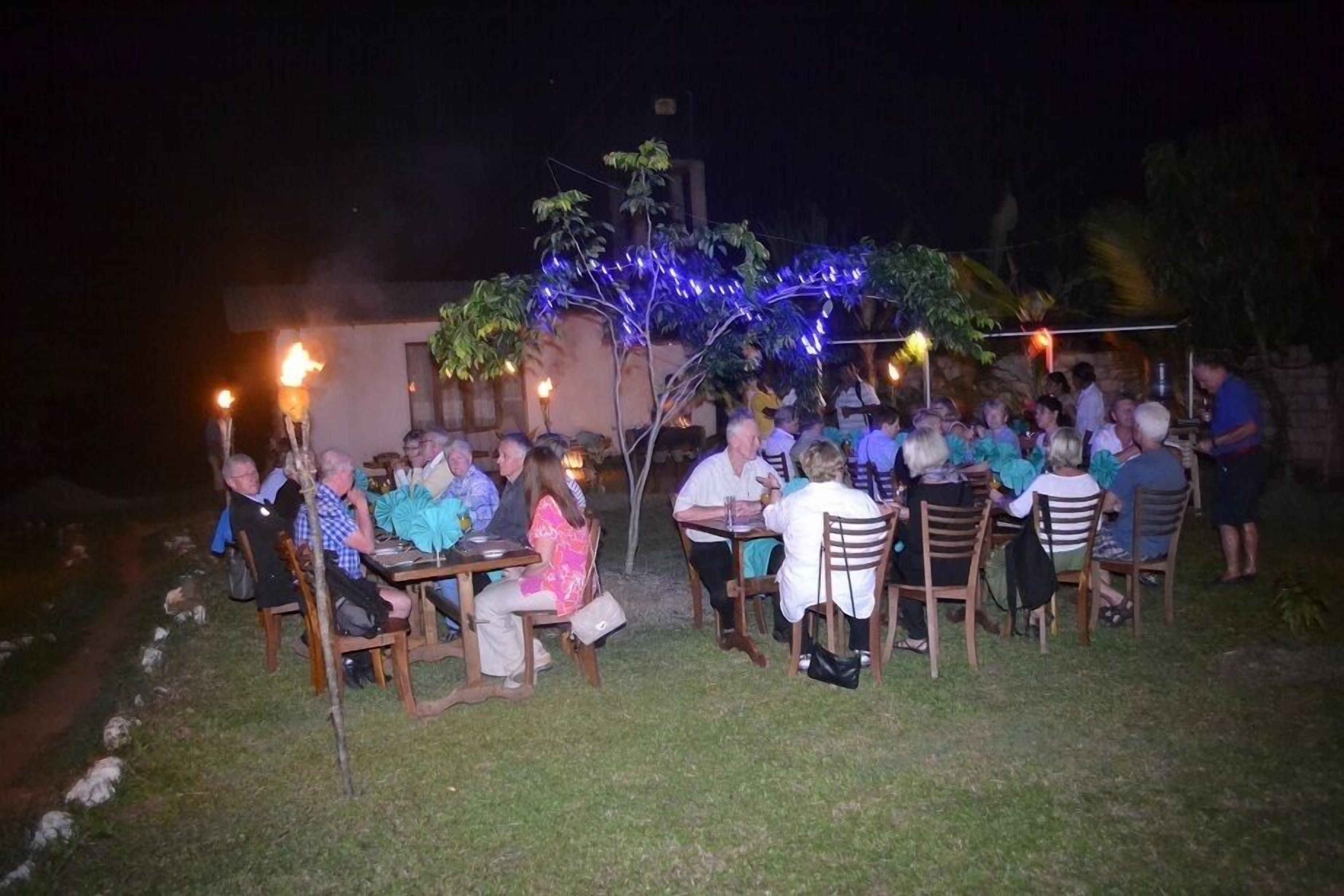 An outdoor dining area with wooden tables and chairs, surrounded by greenery and decorative lights.