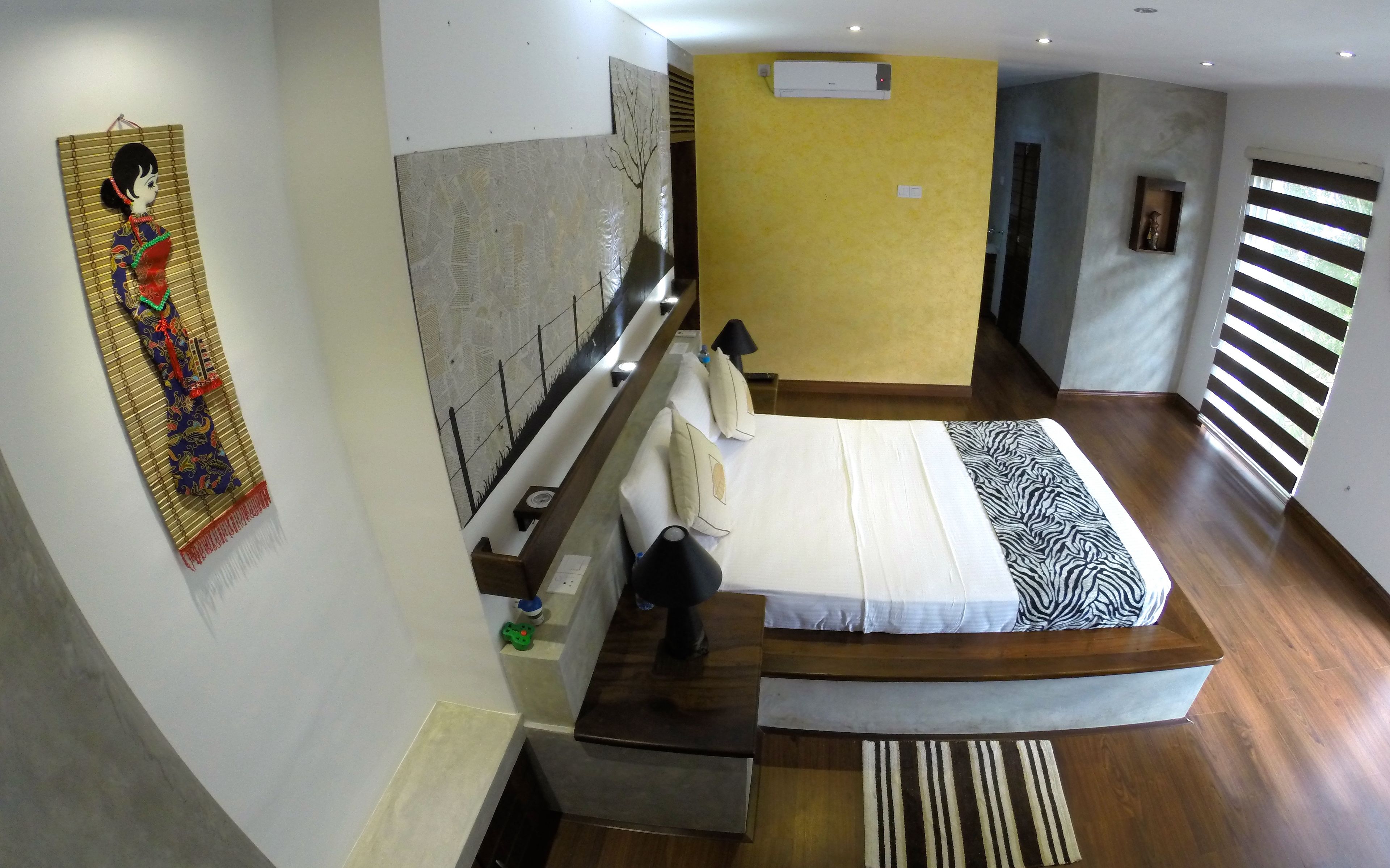 Une chambre d’hôtel moderne avec un grand lit, un bureau en bois, une petite table, une chaise, une table de chevet, un climatiseur mural, une fenêtre avec des stores et un élément décoratif mural.