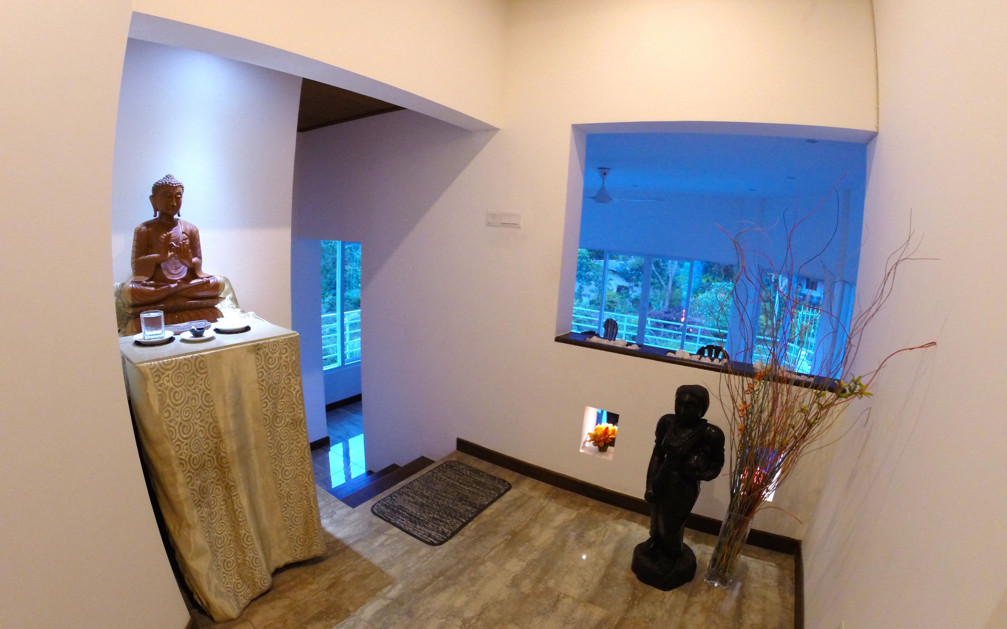 A hallway with a Buddha statue, a window with a view, and a vase with branches.