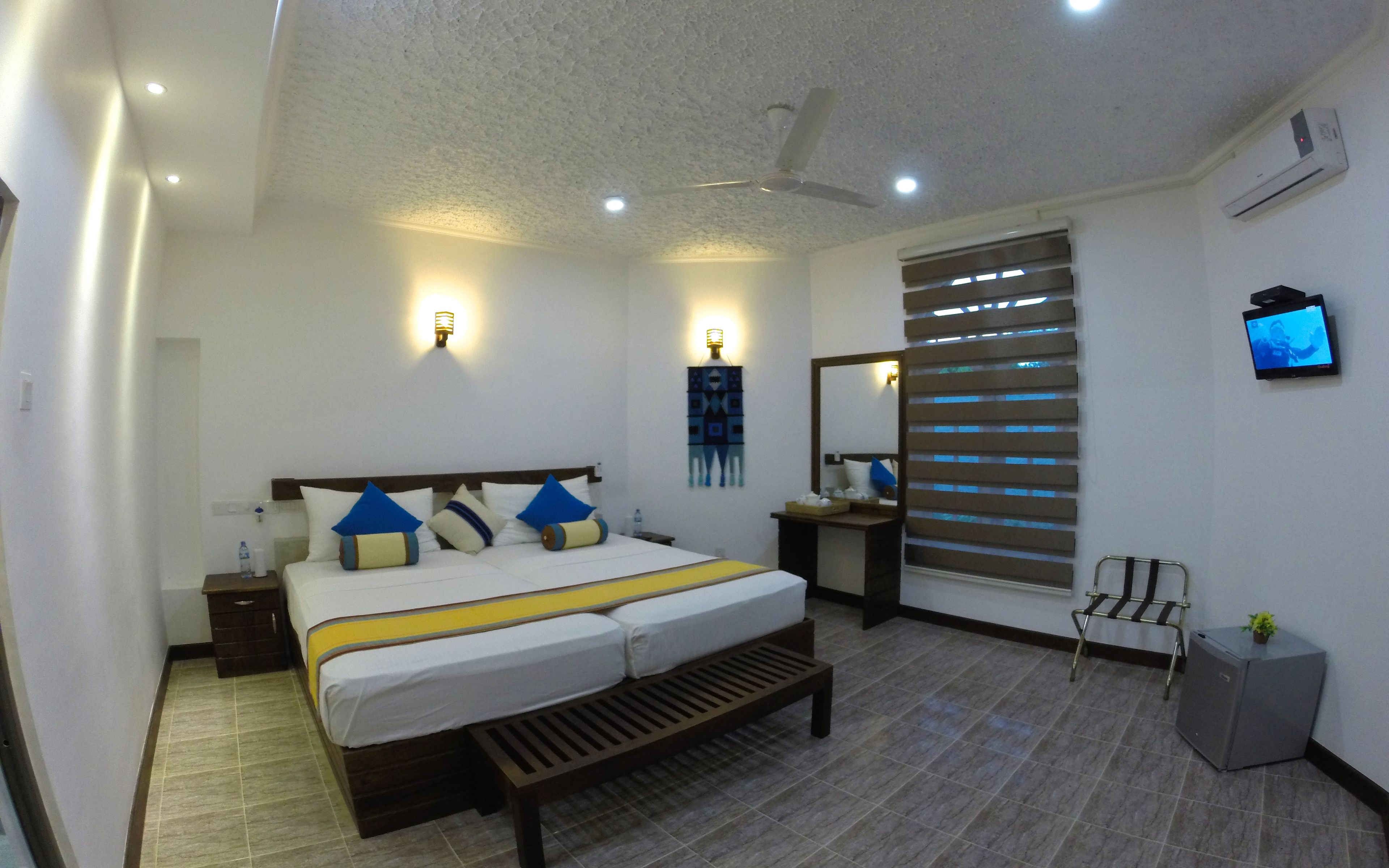 Quarto de hotel moderno com cama, mesas de cabeceira, televisão e ventoinha no teto.