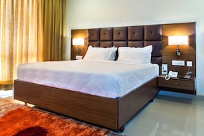 A hotel room with a large bed, two bedside lamps, a nightstand with a phone and a lamp, and a red rug.