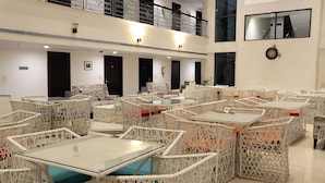 A spacious indoor area with multiple wicker tables and chairs arranged in clusters, a high ceiling with a balcony, and a clock on the wall. - Hotel Saptagiri (Hyderabad)