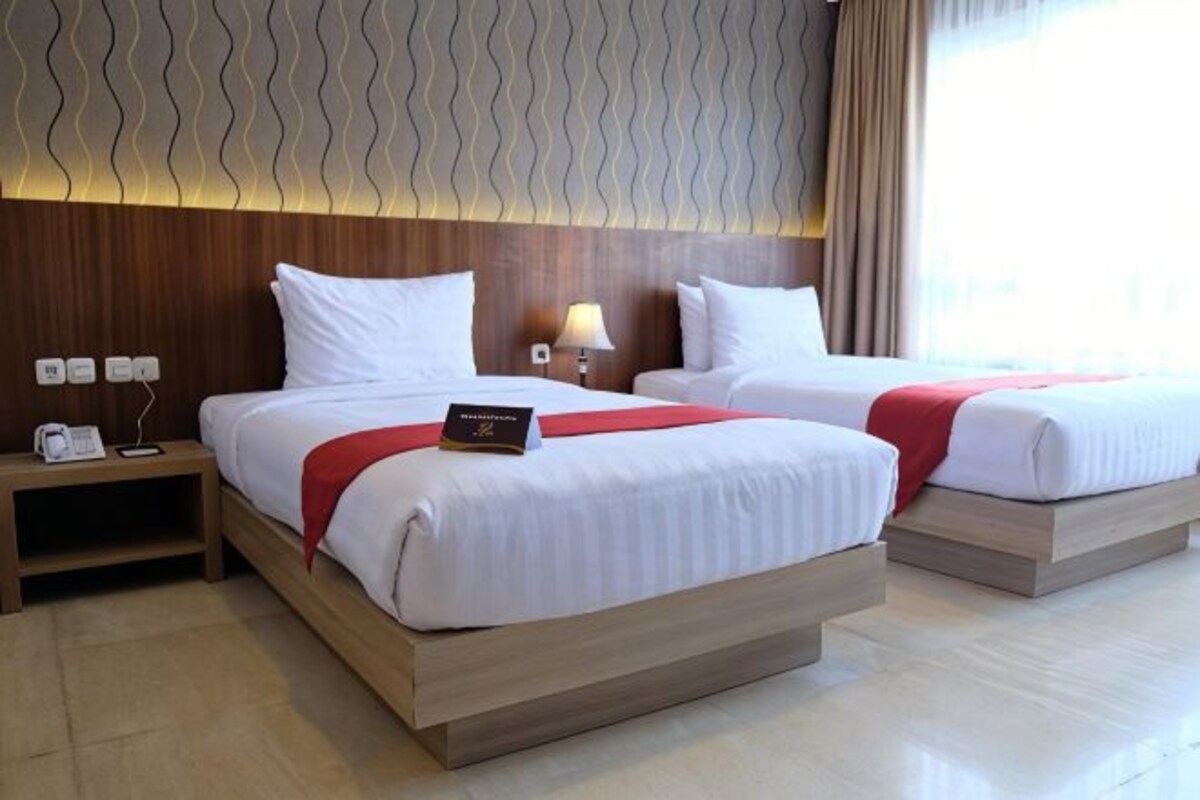 Habitación de hotel con dos camas, una mesita de noche, una lámpara y vistas al exterior.