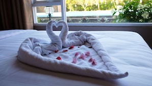 A neatly made bed with a heart-shaped towel arrangement and scattered rose petals. - Mexolie Hotel (Kebumen)