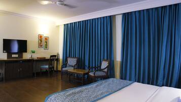 A hotel room with a bed, a desk, two chairs, a television, and a large window with blue curtains.